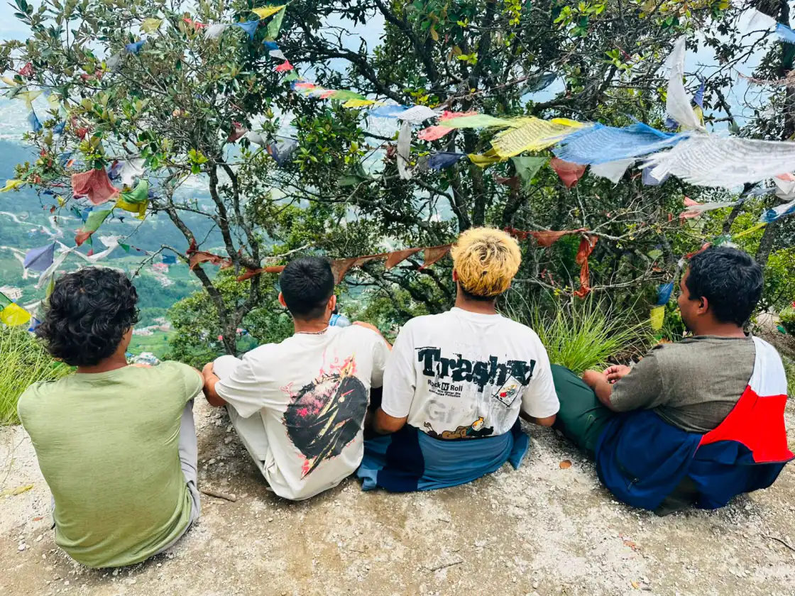 The NepalHRM team resting on a hilltop above the Kathmandu Valley during a team hike, with Buddhist prayer flags strung through the trees